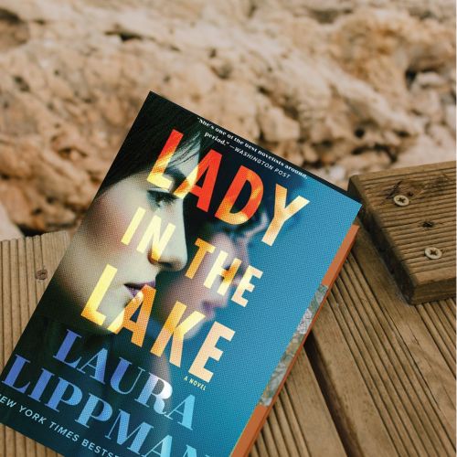 The book Lady in the Lake by Laura Lippman sitting on a wooden deck with sand in the background