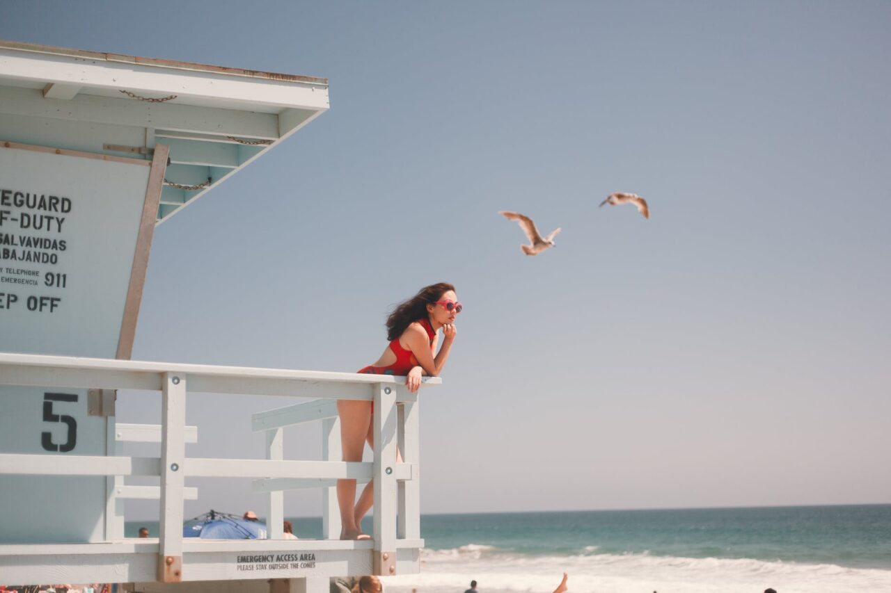 Photo of a woman in a red bathing suit and red sunglasses standing on a lifeguard stand looking out at the ocean