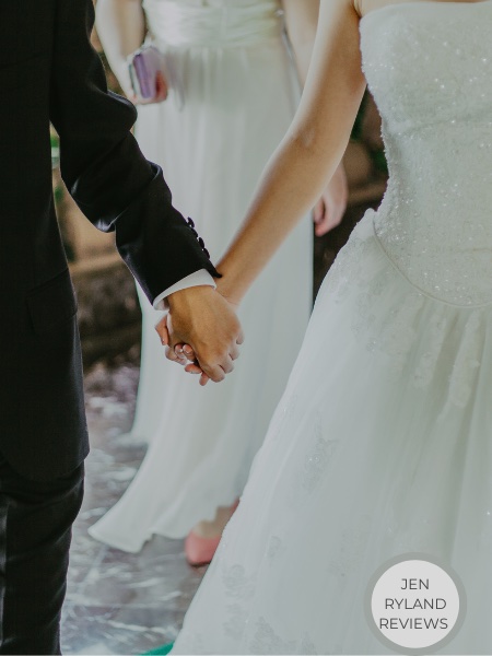 A photo of two people: one in a dark jacket and pants and the other in a white tulle dress. They are holding hands.