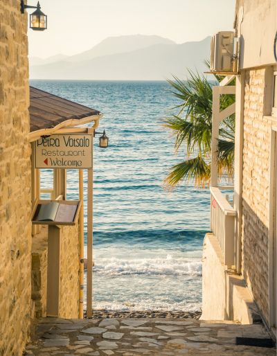 A photo of a street in Greece lined with sun-washed buildings. At the end of the street you can see the ocean.