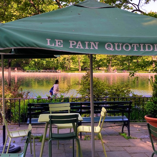 Photo of the cafe at the Conservatory Water in Central Park by Jen Ryland LLC. Photo shows a metal table and charis and green canvas umbrella with a pool of water in the background.