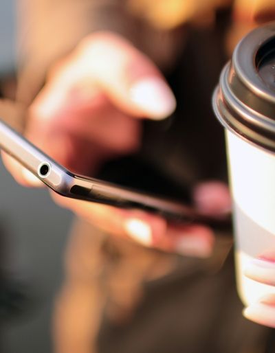 Person holding a cell phone in one hand an a disposable coffee cup in the other hand.