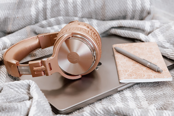 A pair of rose gold headphones sits on top of a pink notebook, a silver laptop, and a gray striped blanket.