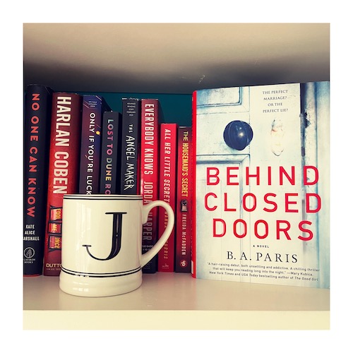 Photo of a hardcover of Behind Closed Doors by B.A. Paris on a shelf of red thrillers with a J mug