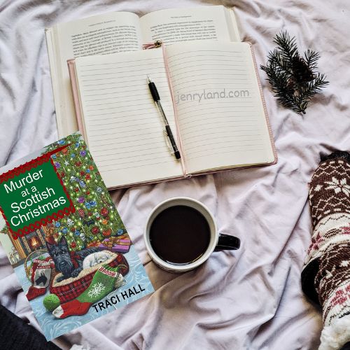 A copy of Murder at a Scottish Christmas on a bed next to a notebook, a cup of coffee, and a foot in a cozy wool sock