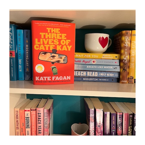 Original photo of a copy of The Three Lives of Cate Kay on a bookshelf next to books and two heart mugs