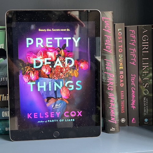 Copy of Pretty Dead Things by Kelsey Coz on a blue shelf with books with pink spines