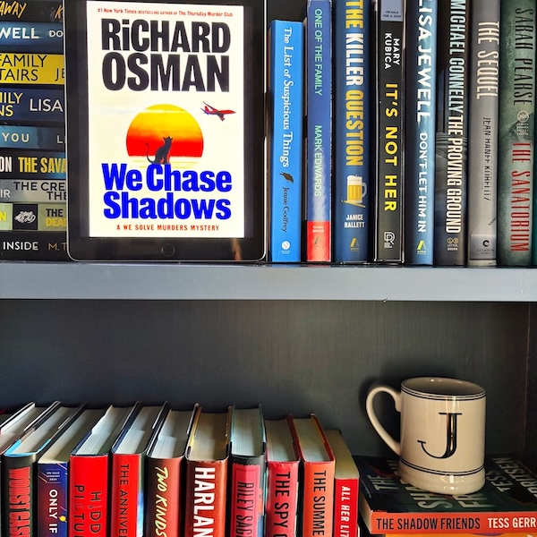 A copy of We Chase Shadows on a bookshelf near other read and blue books and a J mug